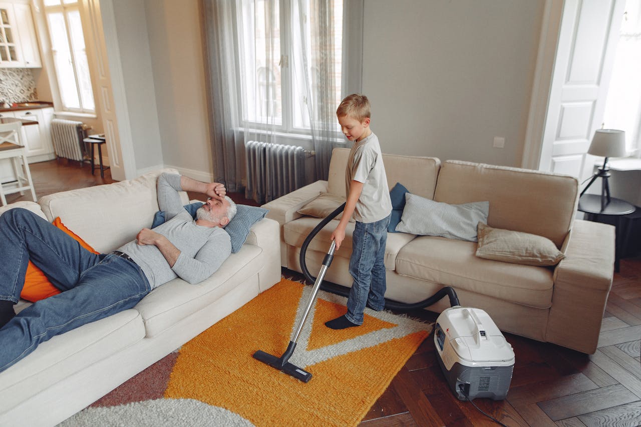 services-01 A young boy vacuums the living room carpet while his grandfather relaxes on the sofa.