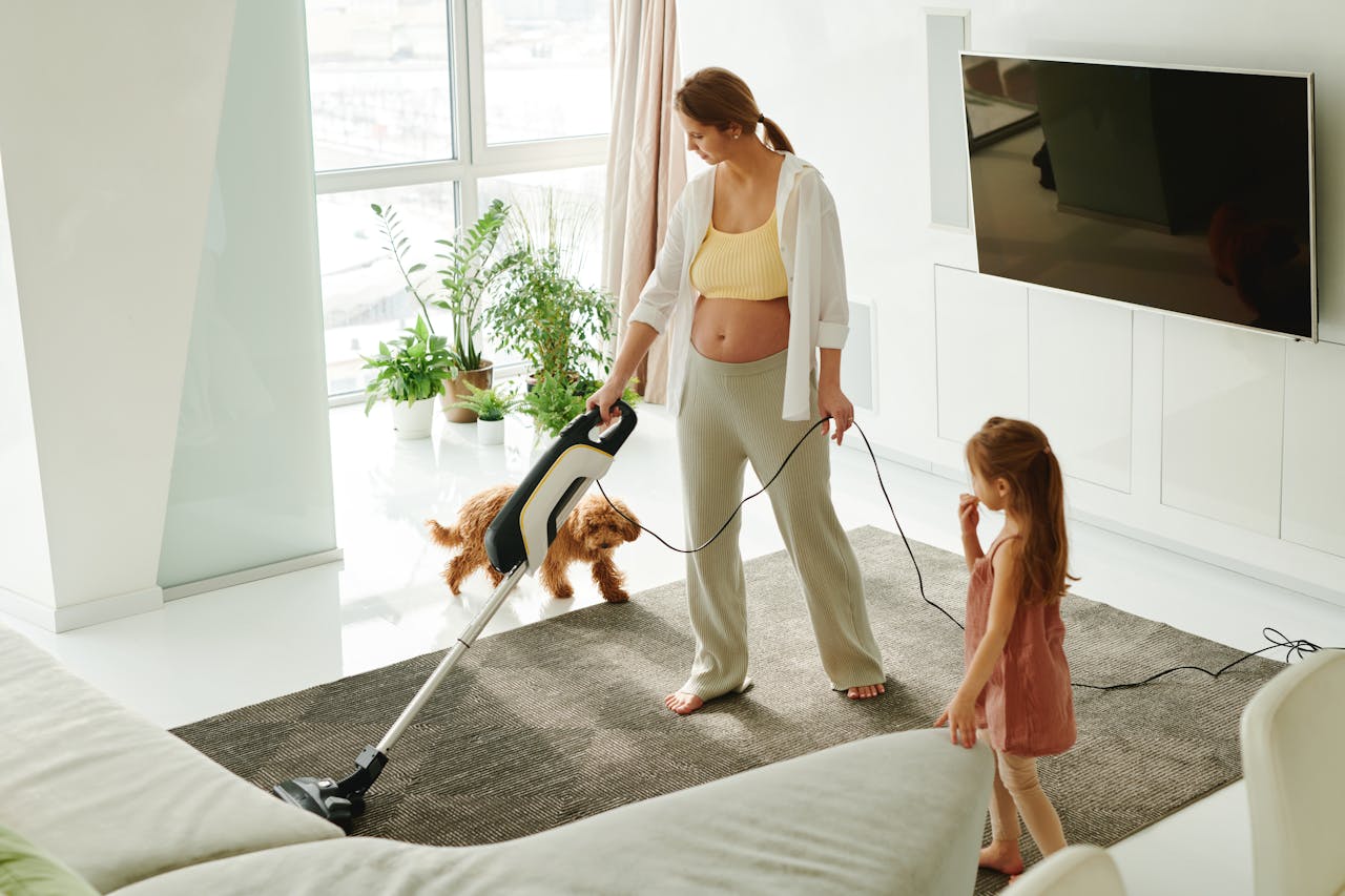 services-02 Pregnant mother cleaning carpet with vacuum as daughter and dog play indoors.